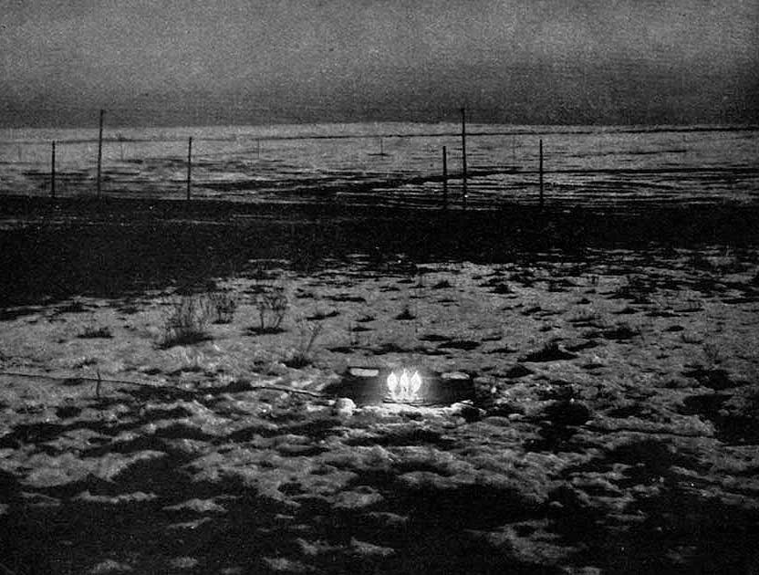 Three incandescent lamps glowing in a hollow in the snow on the open plain at dusk, a line of fence posts beyond